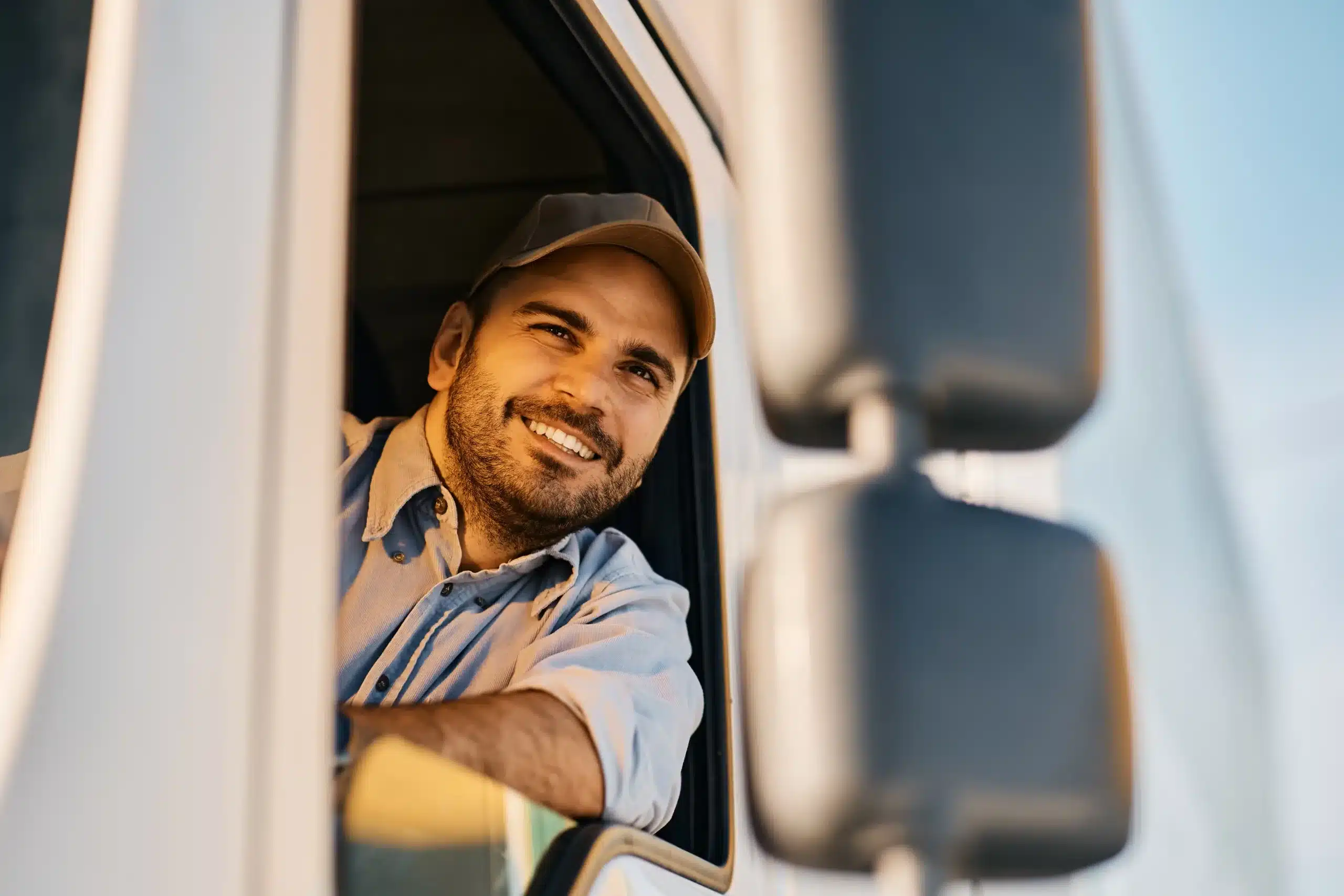 A man wearing a cap and a light shirt smiles while leaning out of the window of a vehicle, possibly a truck, during daylight.