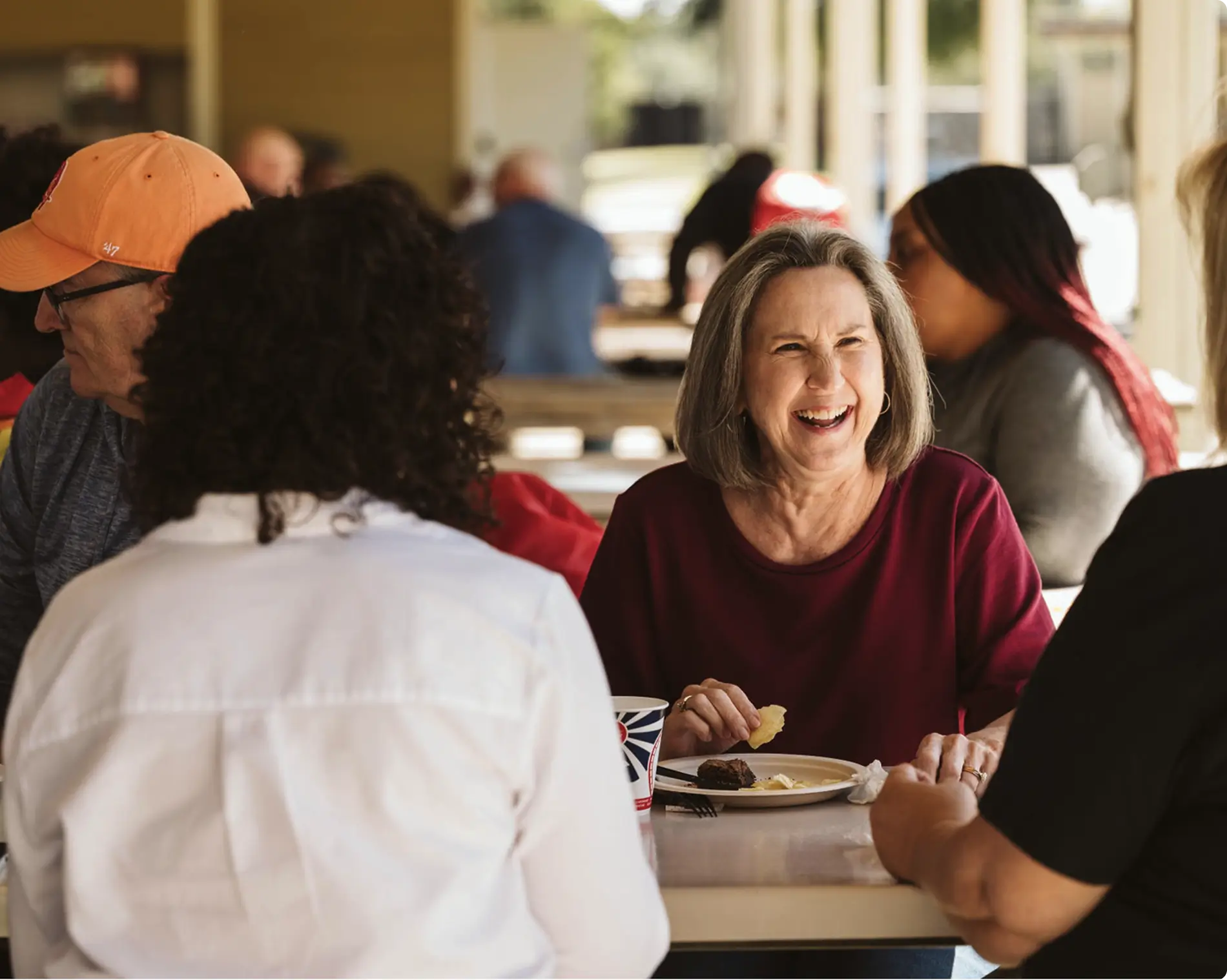 A woman with gray hair laughs while eating with friends at an outdoor table. People are talking and enjoying food in a casual, sunny setting.