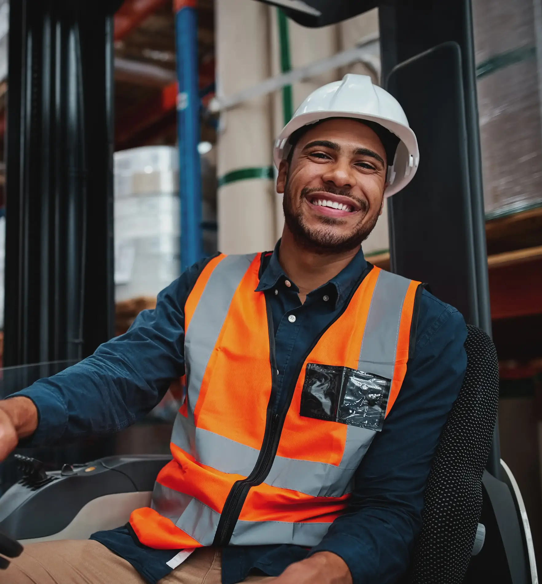 Smiling man wearing a white hard hat and orange safety vest sits in a warehouse setting, looking at the camera.