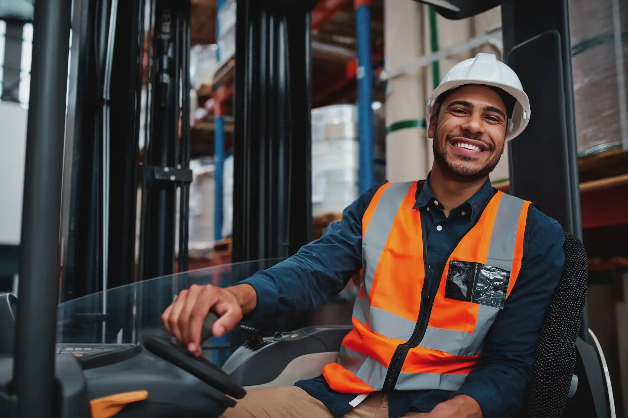 Smiling man wearing a hard hat and orange safety vest sits on a forklift in a warehouse environment.