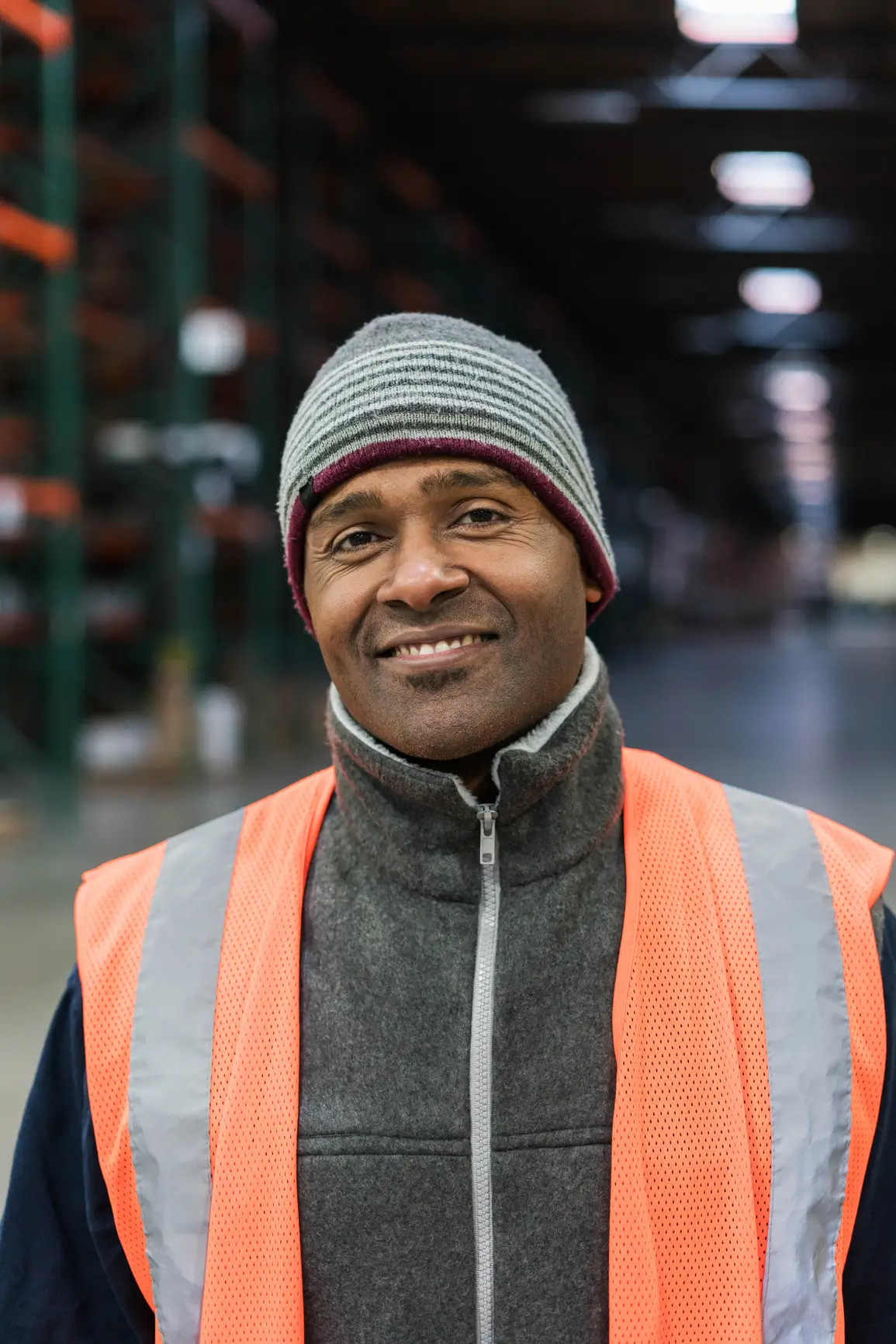 Smiling man wearing a striped beanie and orange safety vest stands in a warehouse with shelves in the background.