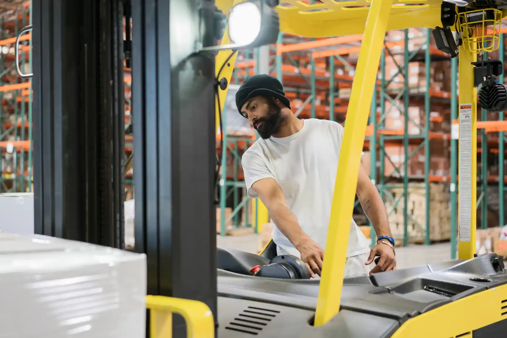 A man in a white t-shirt and beanie operates a yellow forklift in a warehouse with shelves and boxes in the background.