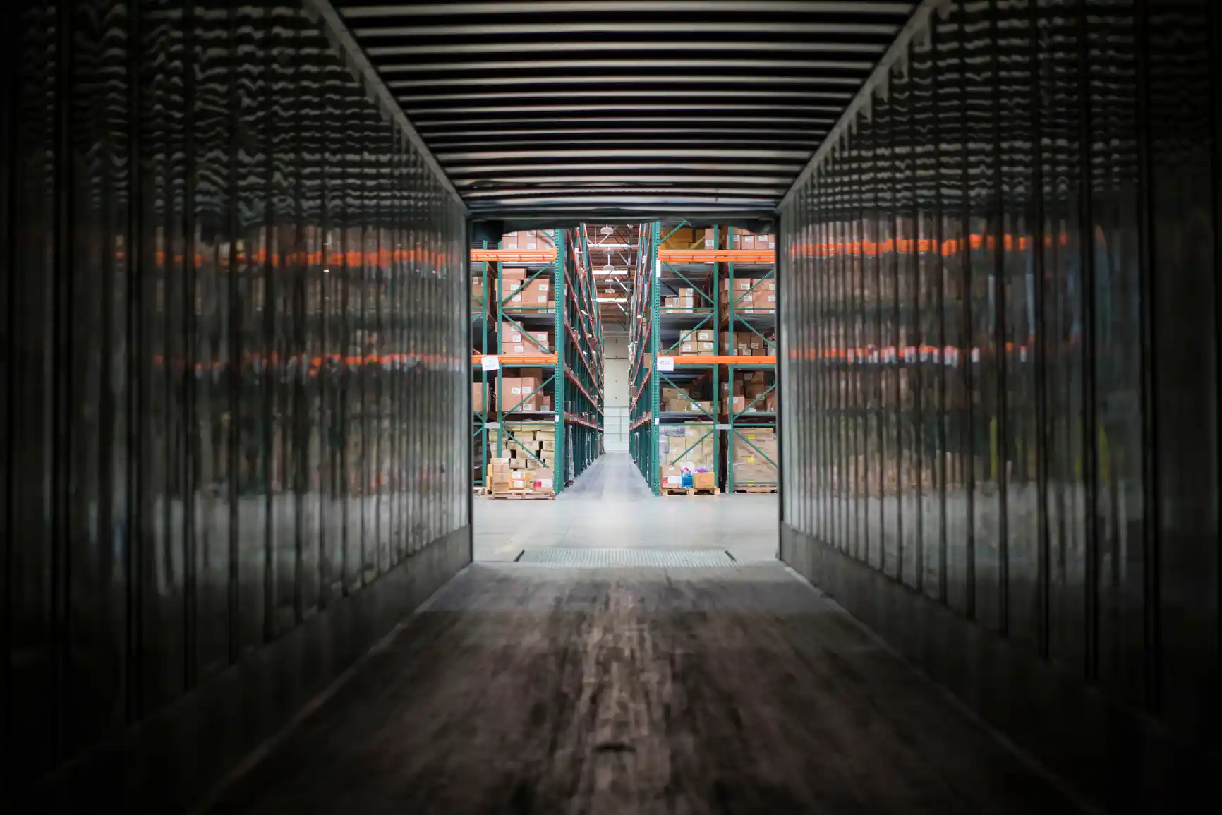 View from inside an empty shipping container, looking out toward industrial warehouse shelving filled with various items.