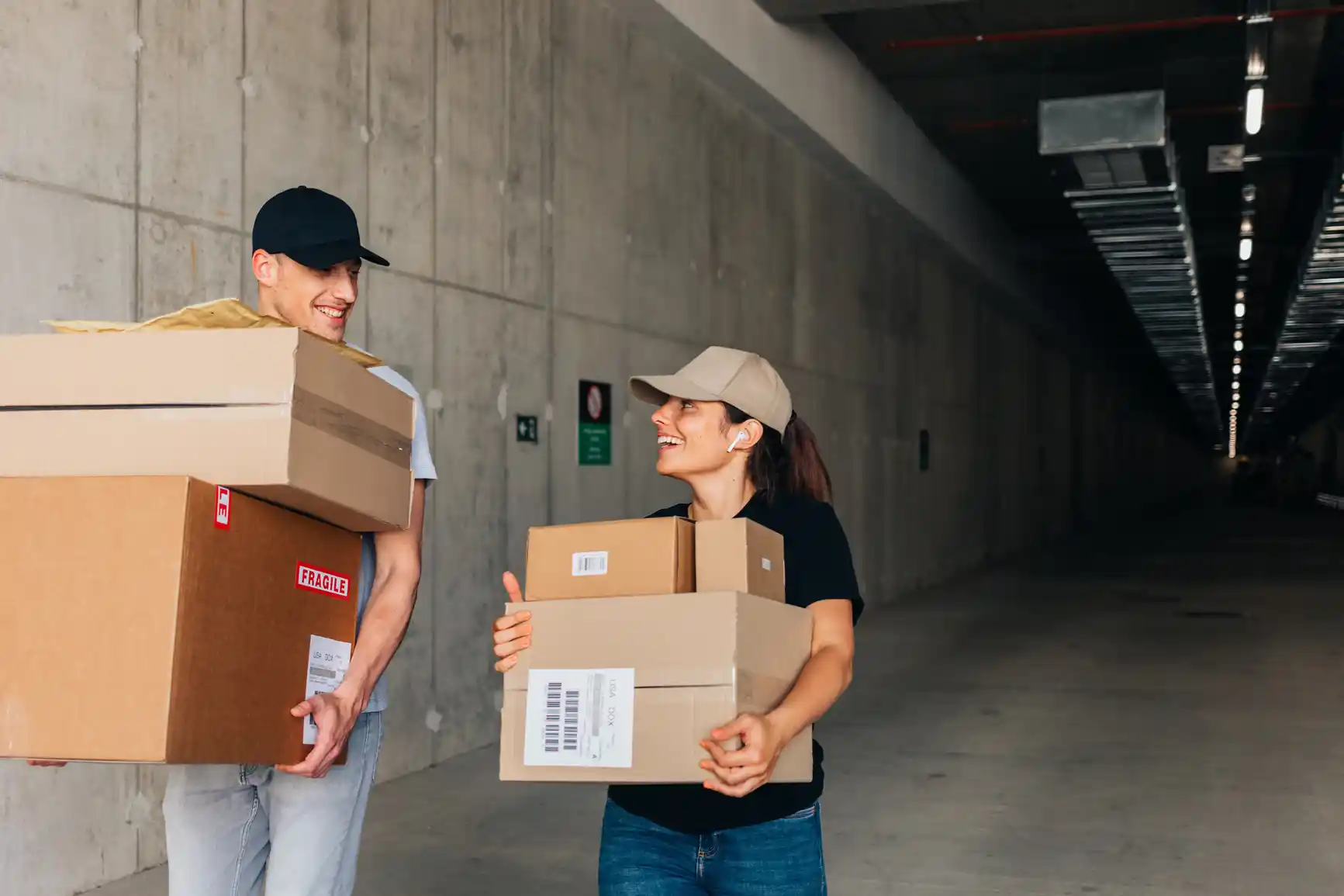 Two people wearing caps smiling at each other while carrying cardboard boxes in a warehouse or industrial hallway.