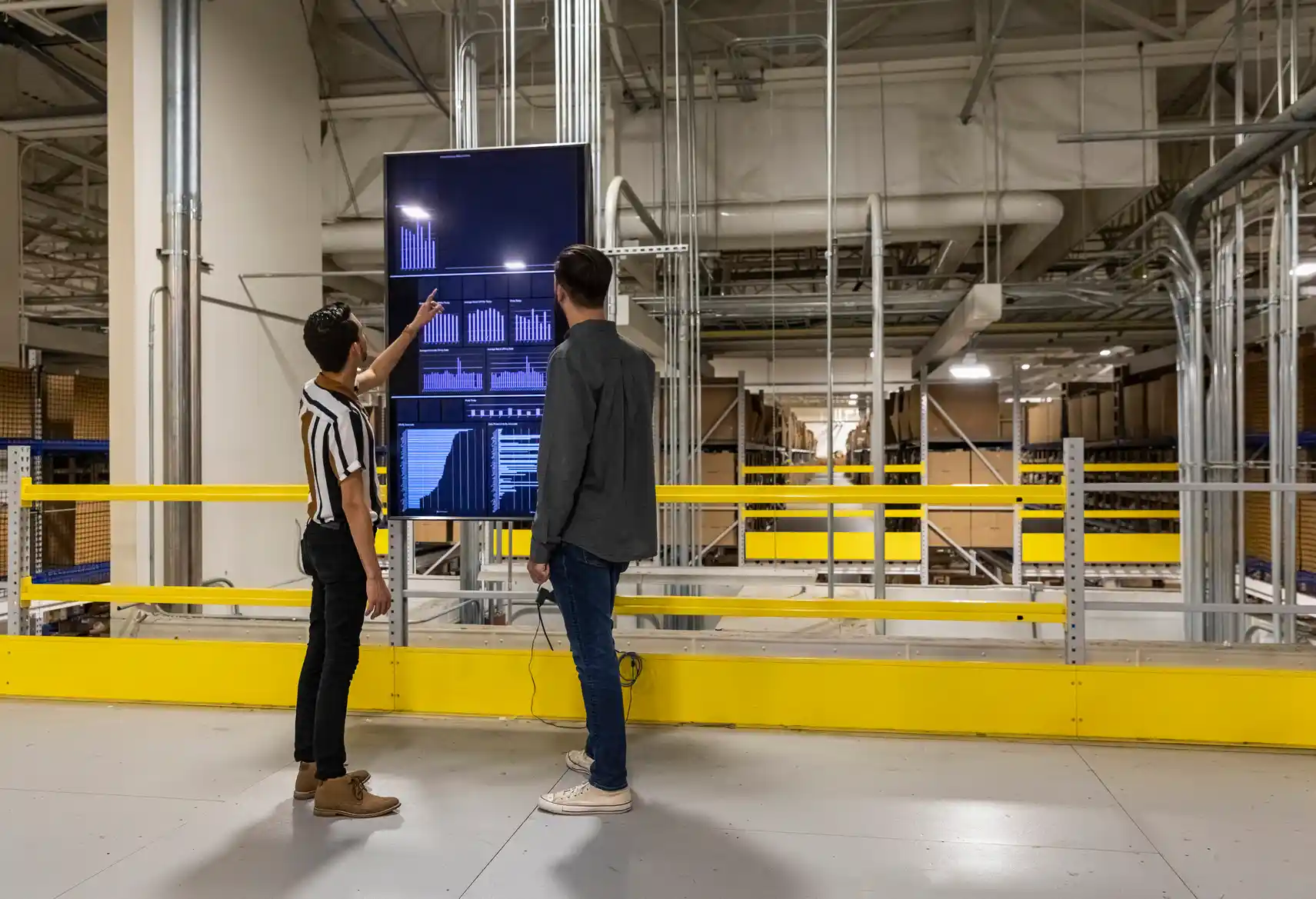 Two people stand in a warehouse, looking at a large screen displaying data and charts. One person points at the screen while the other observes. Industrial pipes and shelves are visible in the background.