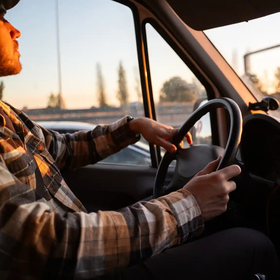 A person wearing a plaid shirt drives a car at sunset, with sunlight streaming through the window and hands on the steering wheel.