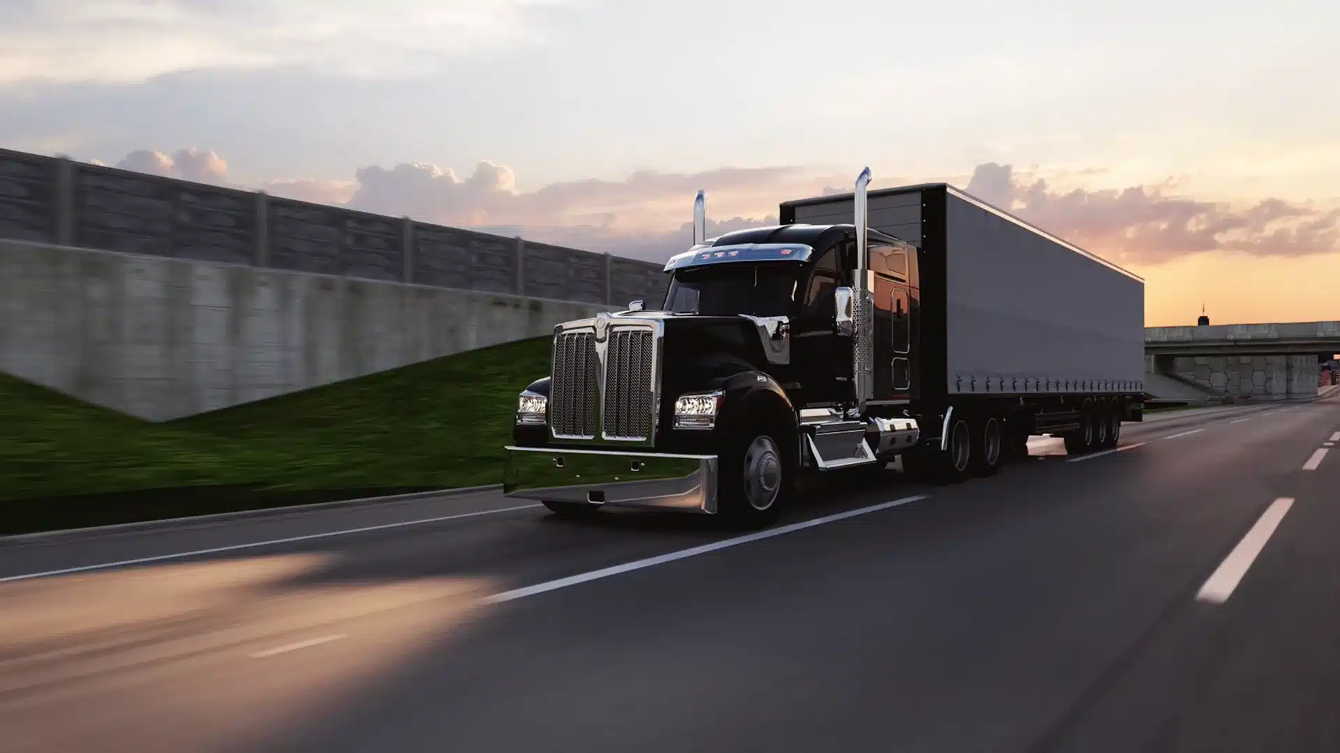 A black semi-truck with a large trailer drives down a highway at sunset, passing a concrete wall and grassy embankment.