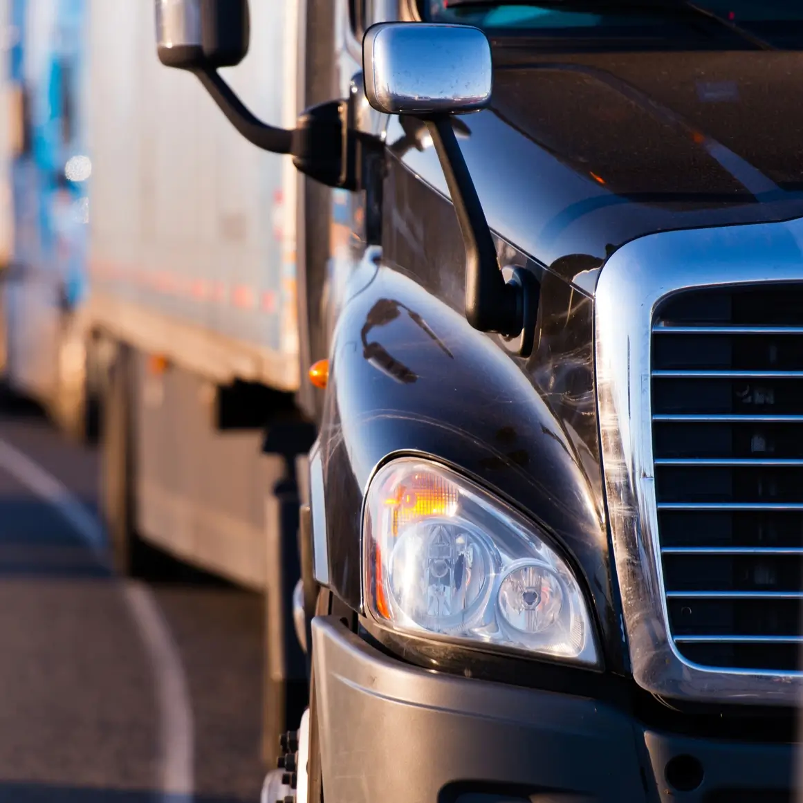 Close-up of the front of a black semi-truck on a road, with another truck visible in the blurred background.