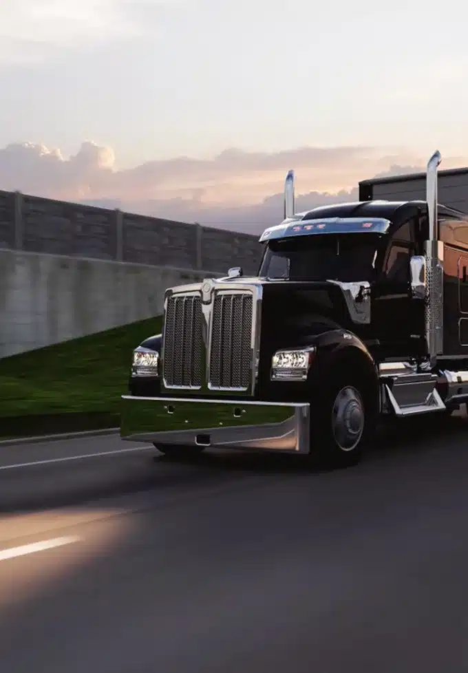 A black semi-truck drives on a highway at sunset, passing a concrete barrier and grassy area, with a cloudy sky in the background.