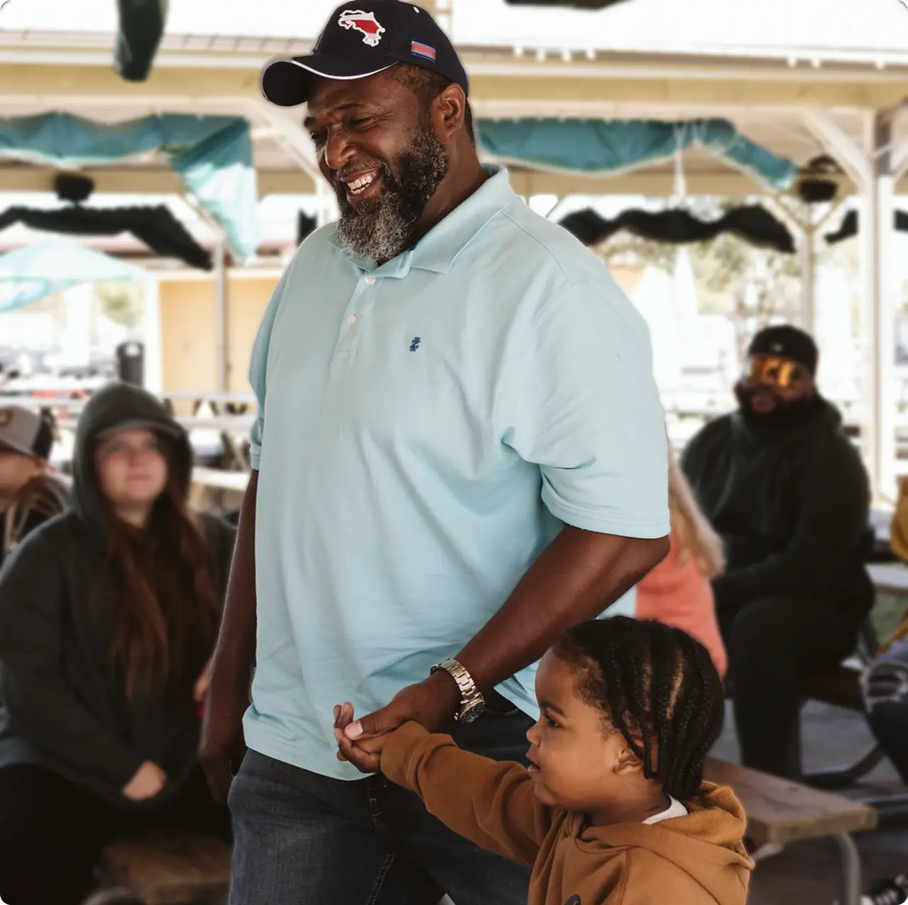 A smiling man in a light blue polo shirt holds hands with a young child in a brown hoodie at an outdoor event with people sitting in the background.