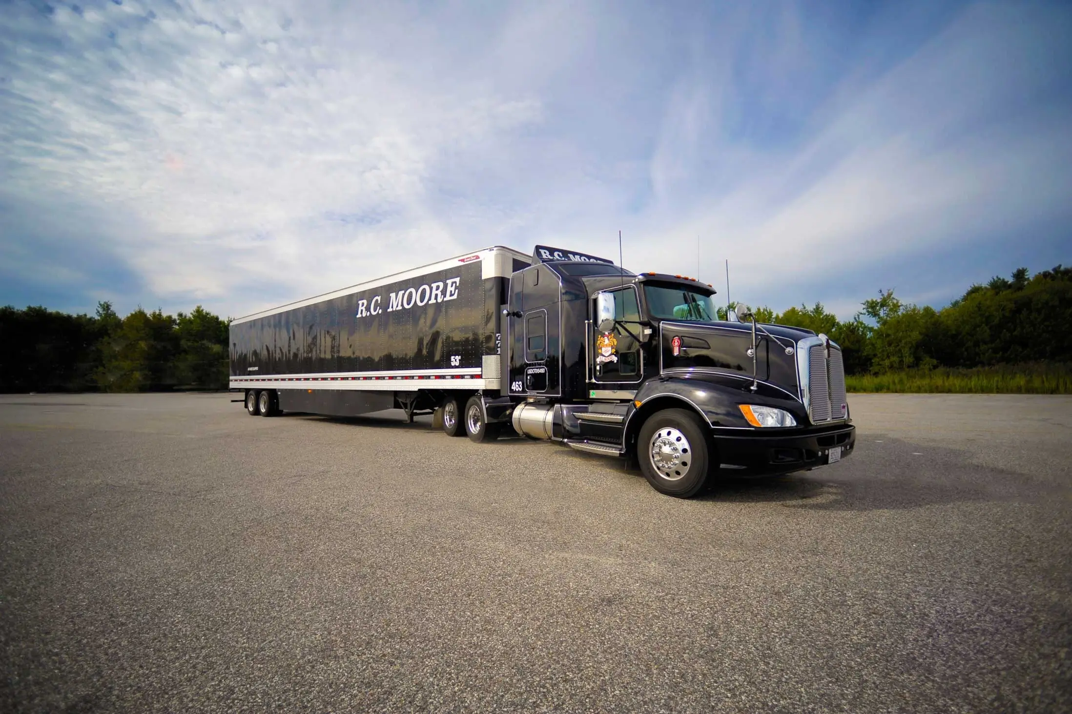 A black semi-truck with R.C. MOORE written on the trailer is parked on a paved lot with trees and a partly cloudy sky in the background.