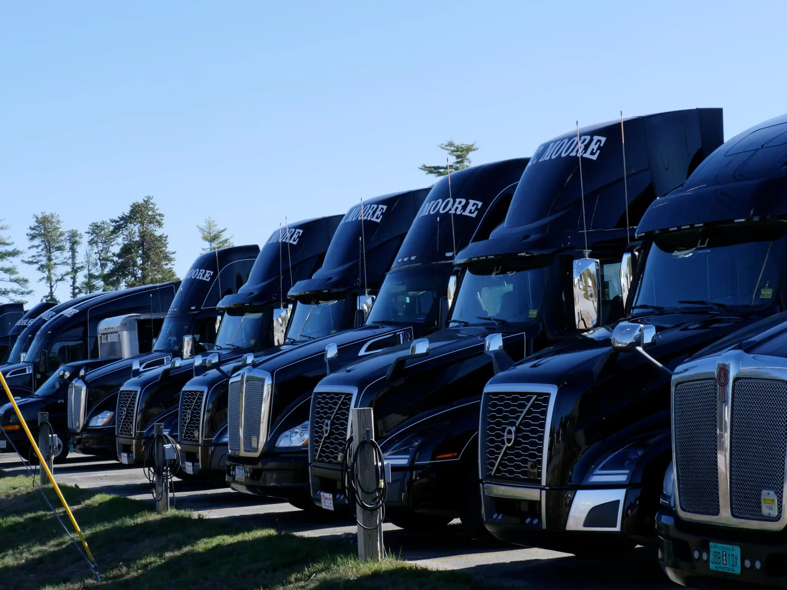 A row of black semi-trucks parked side by side in a lot on a clear, sunny day.